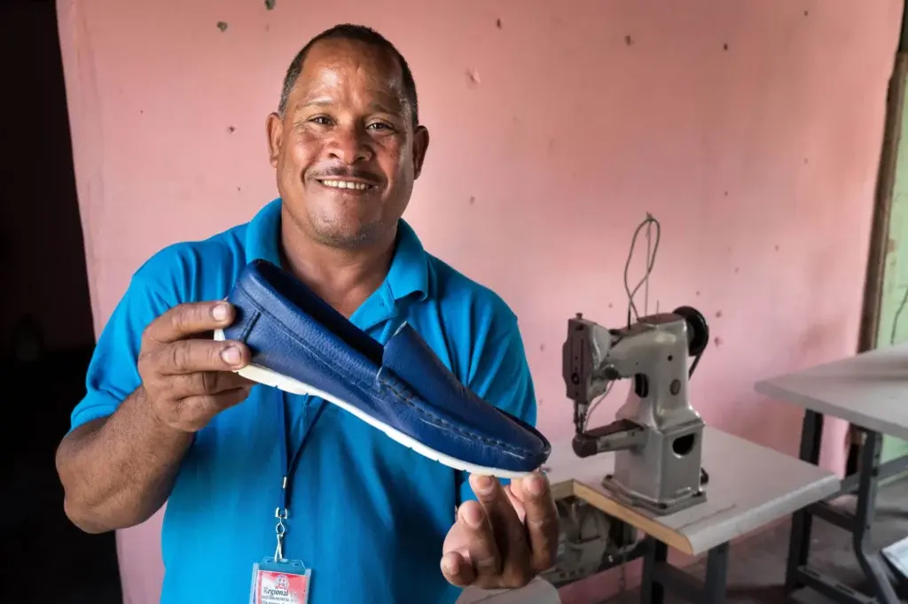 Artesano sonriente mostrando un zapato azul junto a una máquina de coser