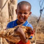 Niño dominicano sonriendo mientras sostiene una gallina, ilustrando los desafíos y adaptaciones de las comunidades rurales frente a las sequías e inundaciones debido al cambio climático en República Dominicana.