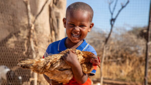 Niño dominicano sonriendo mientras sostiene una gallina, ilustrando los desafíos y adaptaciones de las comunidades rurales frente a las sequías e inundaciones debido al cambio climático en República Dominicana.