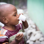 Niño pequeño en República Dominicana bebiendo de un vaso pequeño mientras sostiene un trozo de pan, ilustrando el impacto de la desnutrición y la importancia de una alimentación adecuada en la infancia.