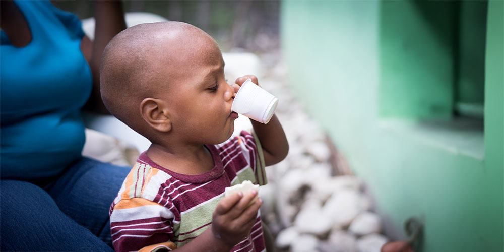 Niño pequeño en República Dominicana bebiendo de un vaso pequeño mientras sostiene un trozo de pan, ilustrando el impacto de la desnutrición y la importancia de una alimentación adecuada en la infancia.