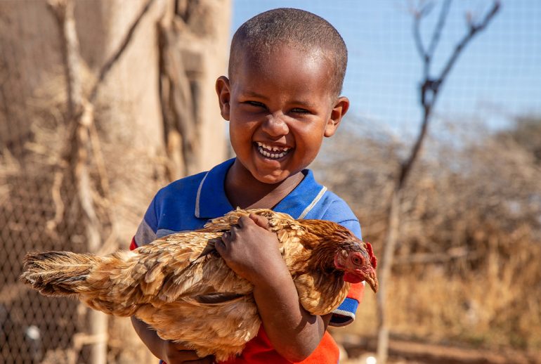 Niño dominicano sonriendo mientras sostiene una gallina, ilustrando los desafíos y adaptaciones de las comunidades rurales frente a las sequías e inundaciones debido al cambio climático en República Dominicana.