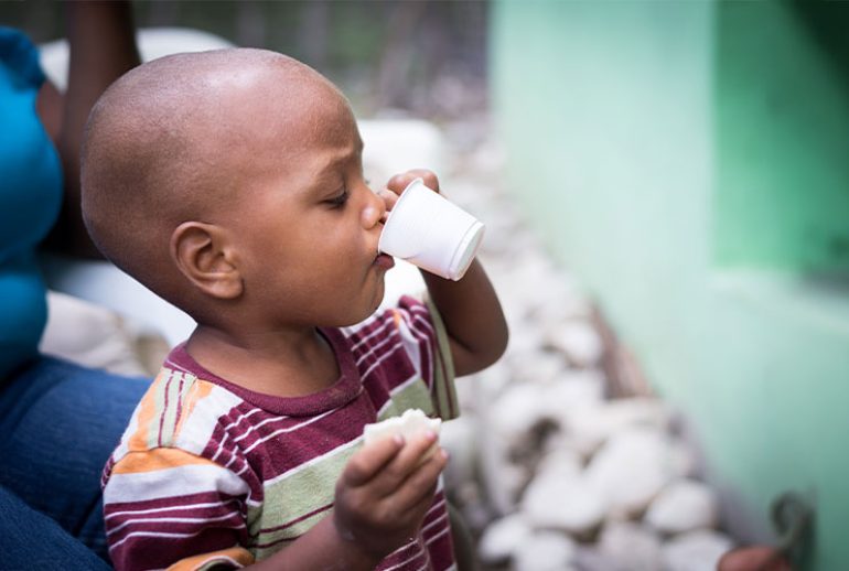 Niño pequeño en República Dominicana bebiendo de un vaso pequeño mientras sostiene un trozo de pan, ilustrando el impacto de la desnutrición y la importancia de una alimentación adecuada en la infancia.